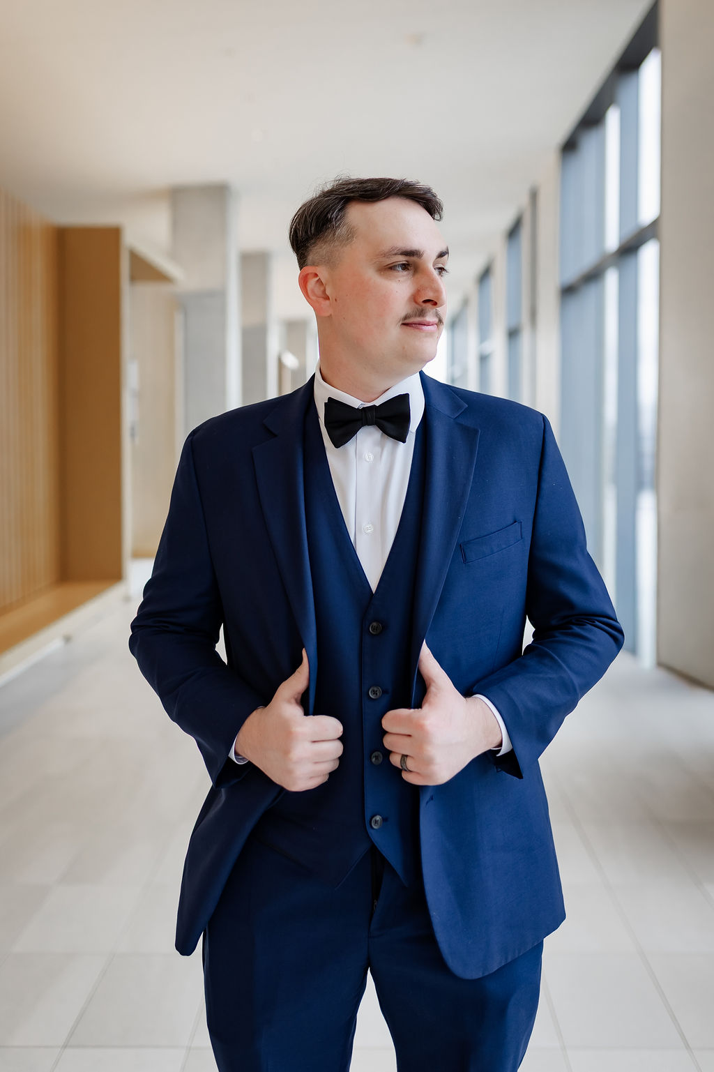 Groom in a navy three piece suit and bow tie in a bright hallway